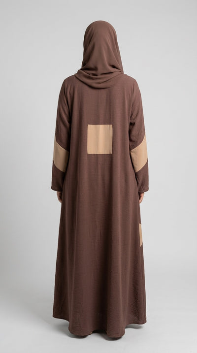 A person shown from the back wearing a long brown abaya or robe with a matching head covering. The garment has a large tan square patch on the back and tan panels on the sleeves, photographed against a plain light gray studio background.