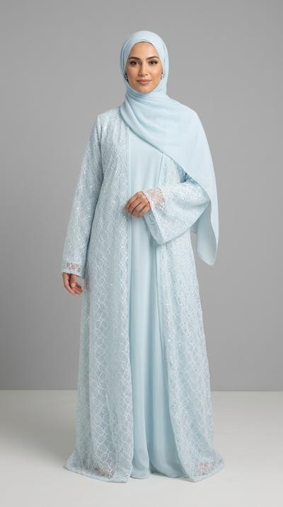A woman wearing a light blue lace abaya with a matching hijab, standing against a neutral gray studio background.