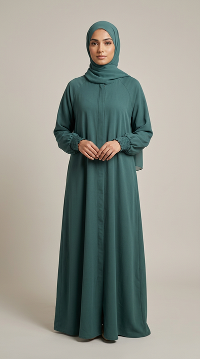 A woman wearing a long-sleeved, floor-length teal abaya with a matching hijab, standing against a neutral beige studio background.