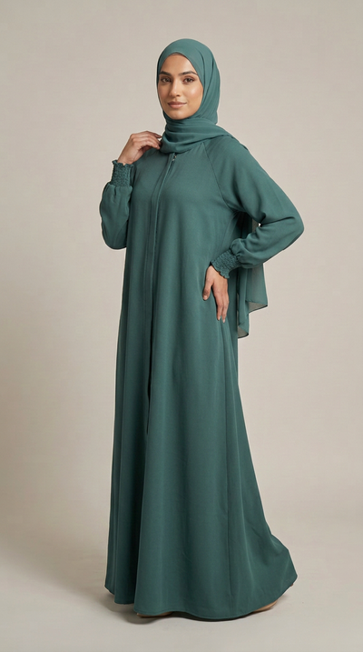 A woman modeling a long-sleeved teal green modest dress with a matching hijab, standing against a neutral beige studio background.