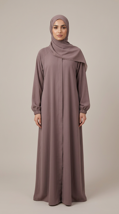 A woman wearing a long, flowing mauve-gray abaya with a matching hijab, standing against a plain beige studio background.