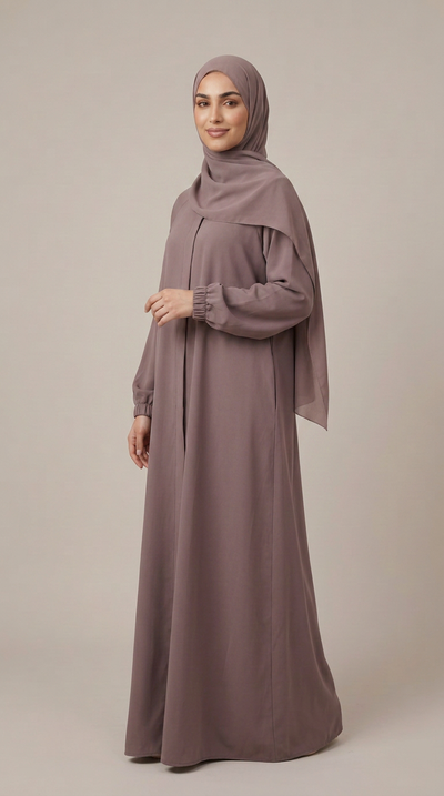 A woman wearing a long, flowing abaya in a muted dusty mauve color, paired with a matching hijab, standing against a solid light beige studio background.