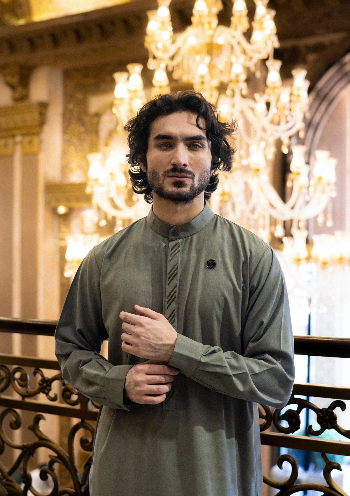 Man in a green traditional outfit standing in front of a large ornate chandelier.