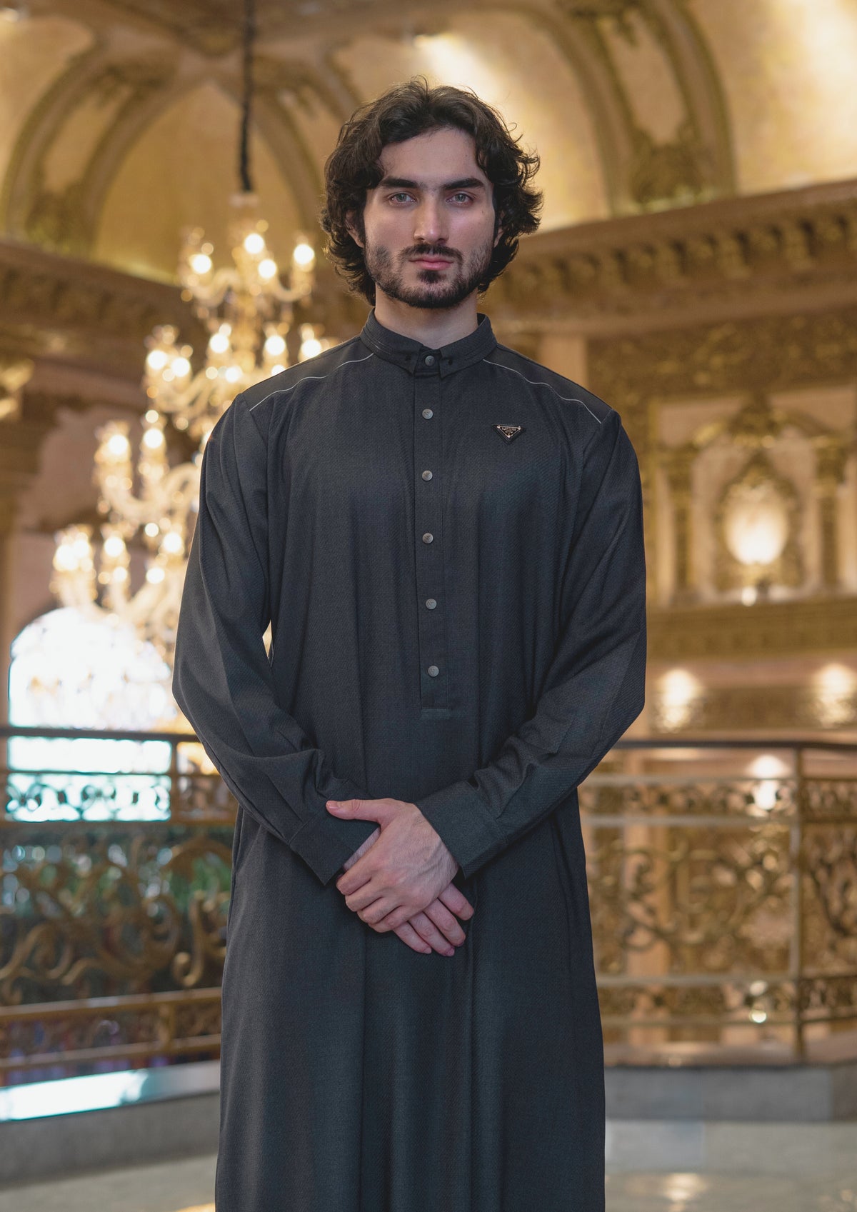 Man wearing a dark traditional outfit standing in an ornate indoor setting with chandeliers.