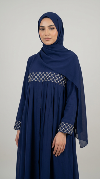 A woman wearing a navy blue modest dress with a matching hijab, featuring a geometric patterned trim at the waist and cuffs, photographed against a neutral light gray background.