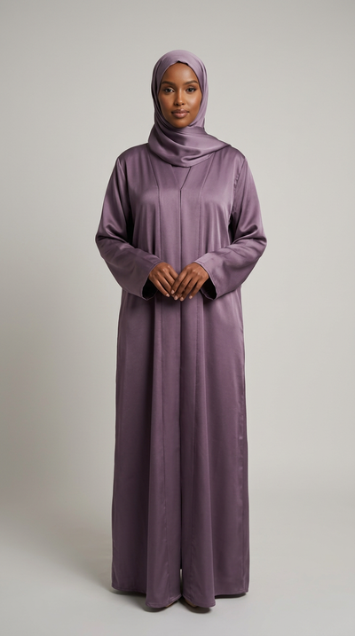 A woman wearing a long-sleeved, floor-length mauve-purple abaya and matching hijab, standing against a neutral light gray studio background.