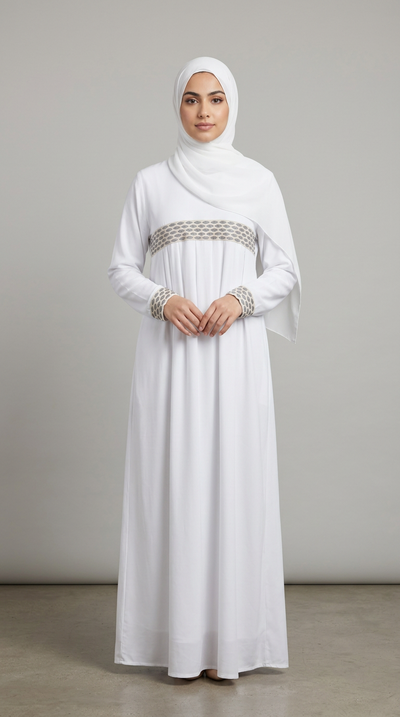 A woman wearing a long white modest dress with a matching hijab, standing against a plain light gray studio background. The dress has a decorative metallic patterned waistband and matching patterned cuffs.