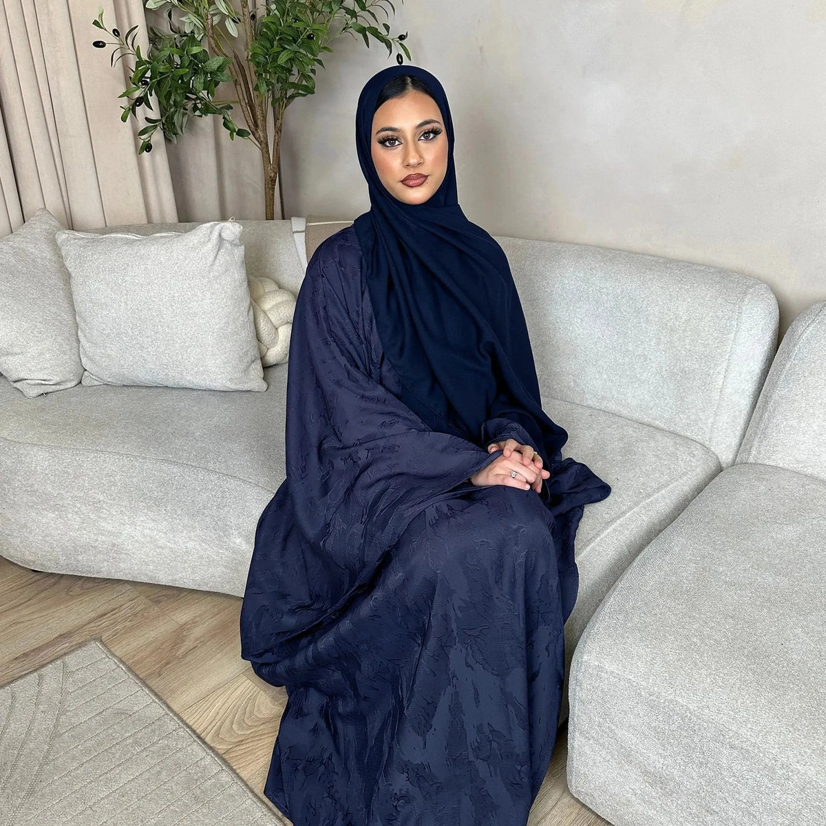 Woman in a navy abaya sitting on a gray sofa in a living room.
